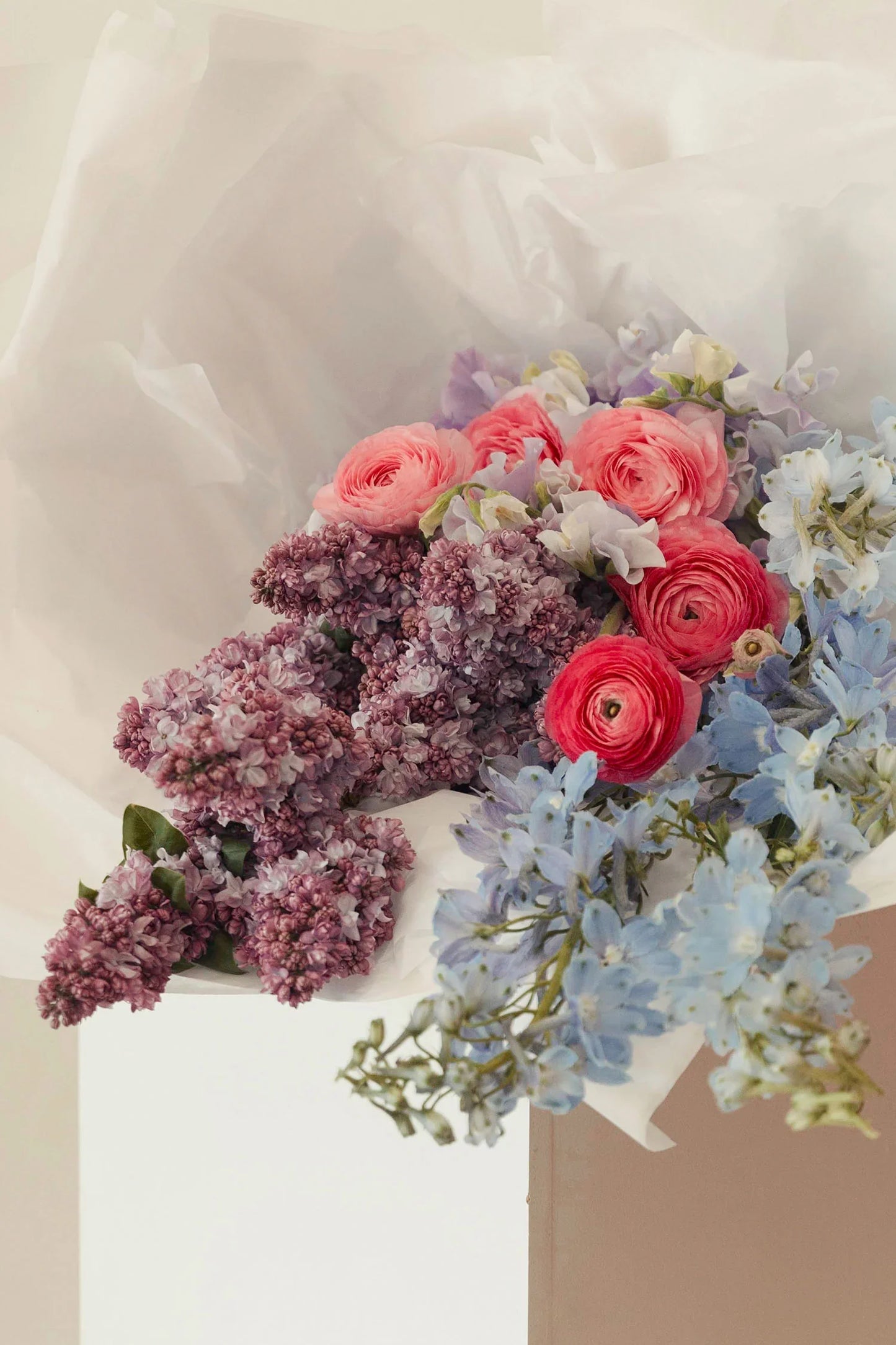 Pastel bouquet with pink ranunculus, lilac flowers, and blue delphiniums wrapped in white paper