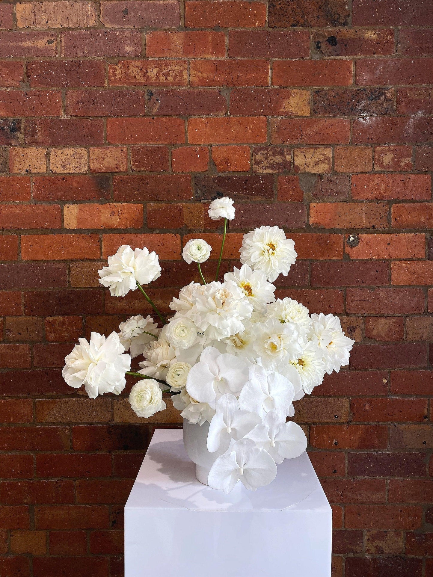 White flower arrangement in ceramic vase on pedestal against red brick wall